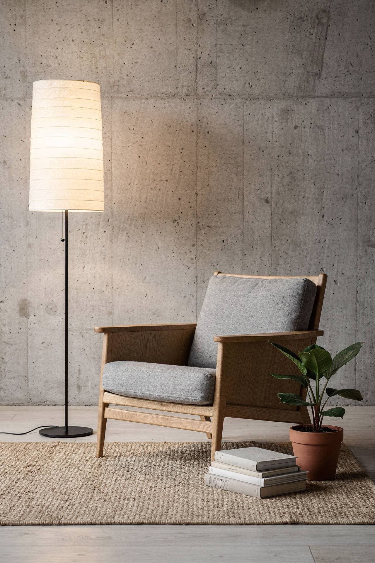 An oak lounge chair with grey wool cushions beside a tall paper floor lamp and a stack of books