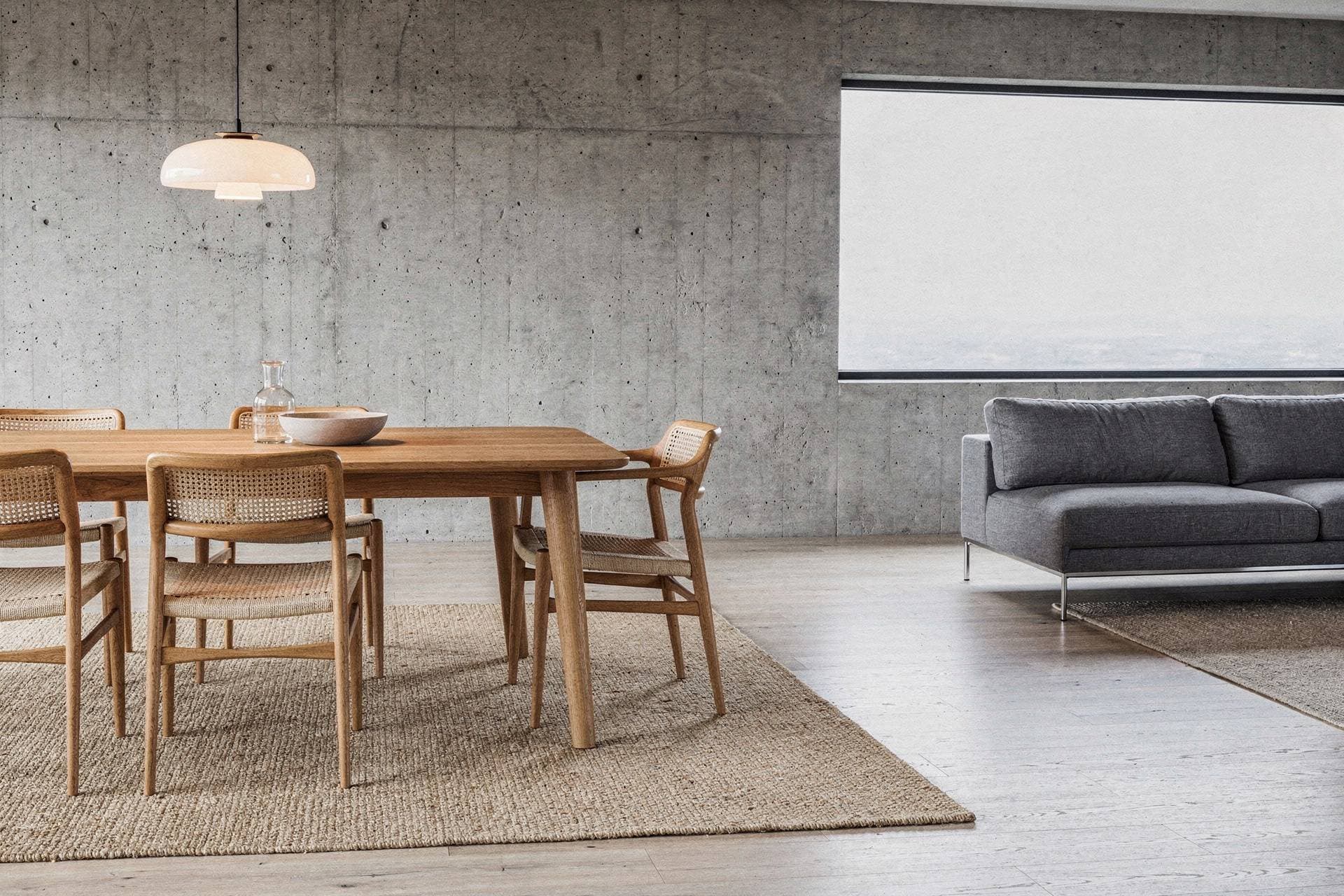 An oak dining table with paper-cord chairs under an opal glass pendant, a grey sofa beyond